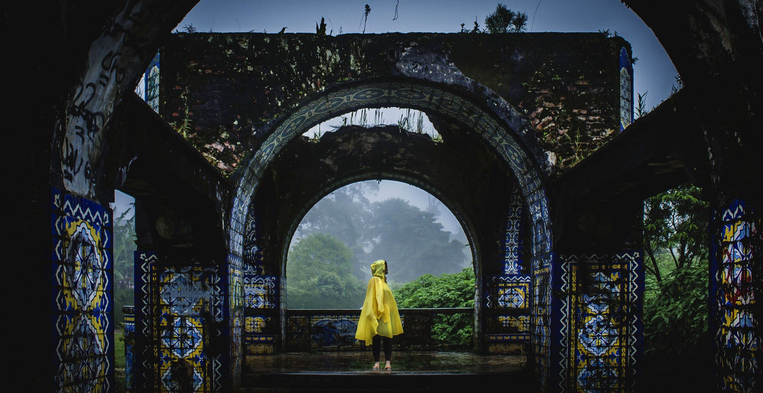 A person wearing a yellow rain poncho stands beneath ornate stone arches overlooking a misty landscape.