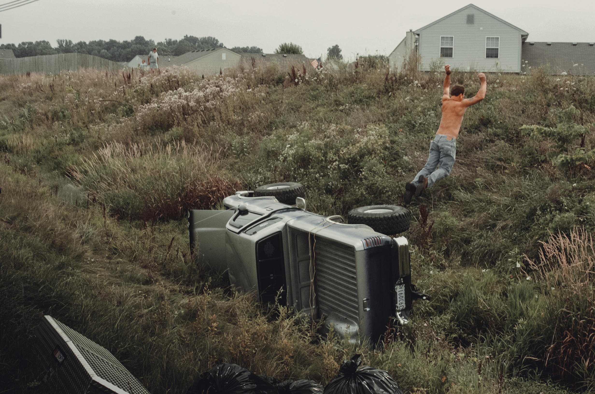 A person leaps away from an overturned vehicle in a grassy field.