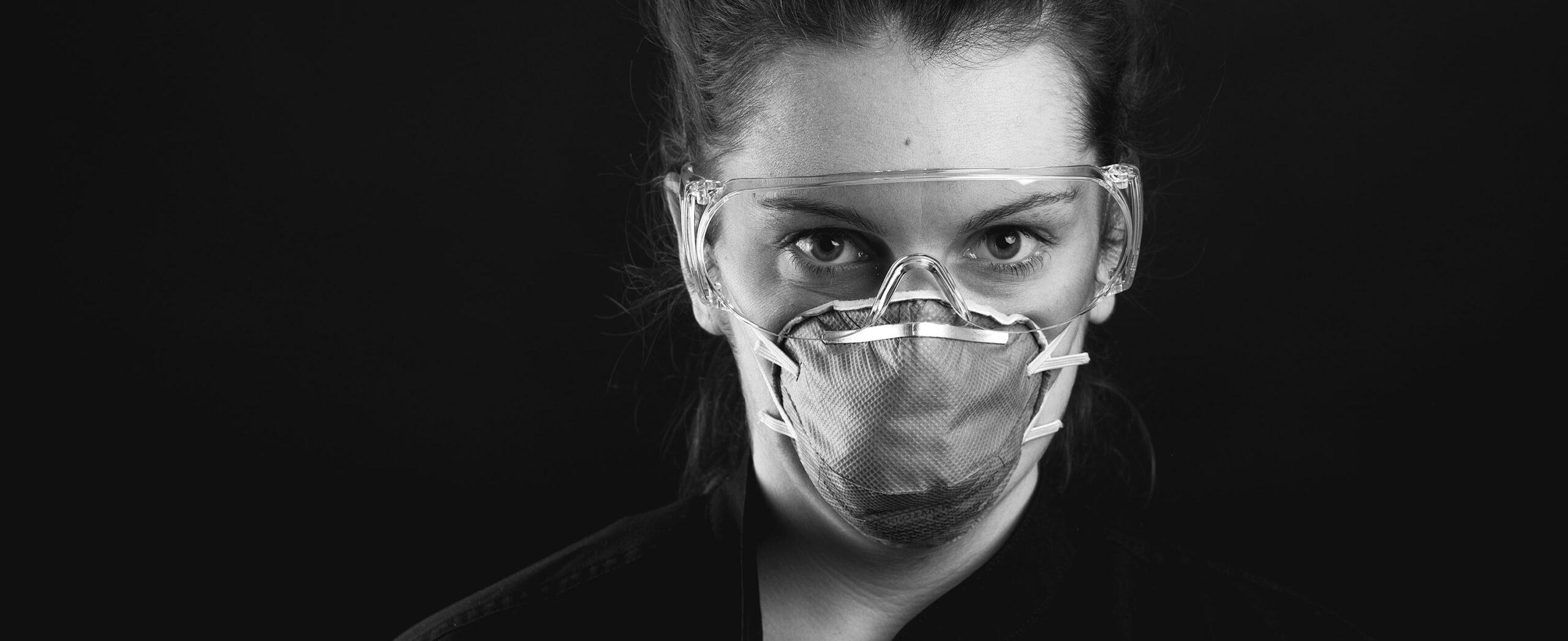 Black-and-white close-up portrait of Caitlyn wearing protective goggles and a respirator mask, looking directly into the camera.