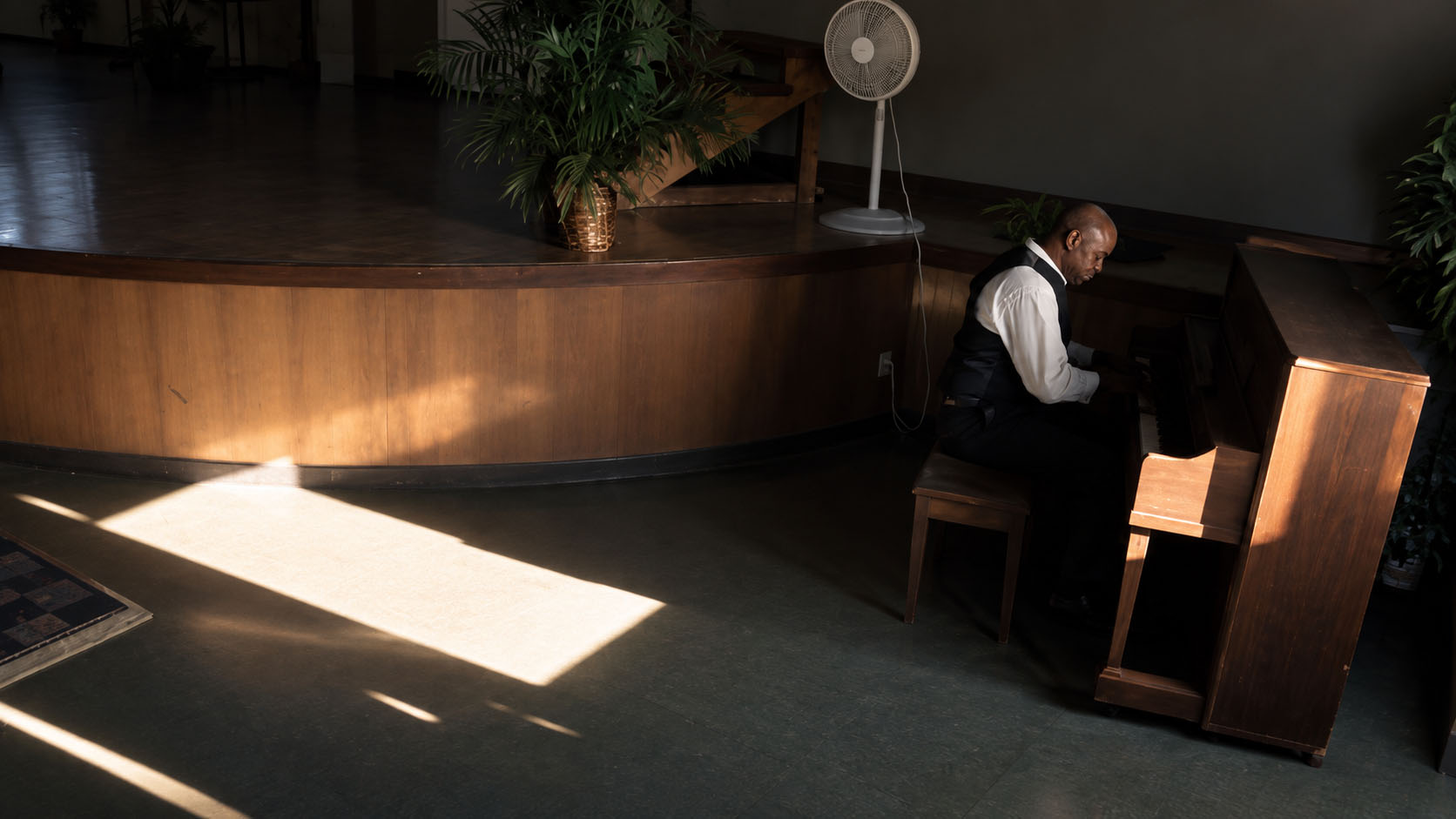 Justin Echols seated at a piano in a dark room, with a bright rectangle of sunlight crossing the floor.