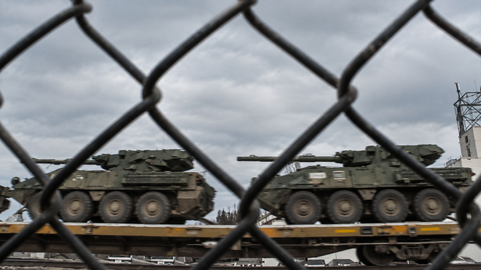 Military vehicles riding on flatbed rail cars, seen through a chain-link fence.